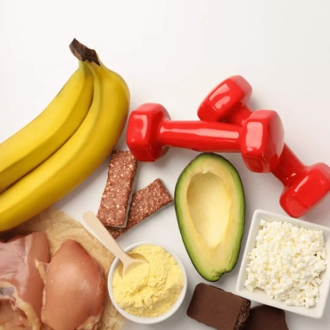 Imagen de dieta nutricional balanceada para realizar deporte de competición o carreras. Se ven dos bananas, dos barritas de cereal, dos pechugas de pollo, queso magro, palta, chocolate en barra y dos pesas livianas para realizar ejercicio.