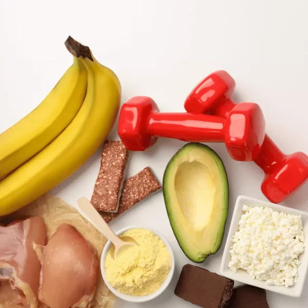 Imagen de dieta nutricional balanceada para realizar deporte de competición o carreras. Se ven dos bananas, dos barritas de cereal, dos pechugas de pollo, queso magro, palta, chocolate en barra y dos pesas livianas para realizar ejercicio.